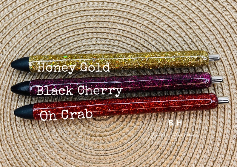 Puede incluir: Tres bol&iacute;grafos de purpurina con tapas negras y detalles plateados. Los bol&iacute;grafos est&aacute;n etiquetados como "Honey Gold", "Black Cherry" y "Oh Crab".