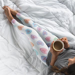 Puede incluir: Una persona lleva puestas unas mallas blancas con un estampado de corazones de caramelo coloridos que dicen "Kiss Me" y "You Rock". La persona está sosteniendo una taza amarilla con un líquido marrón en su interior.