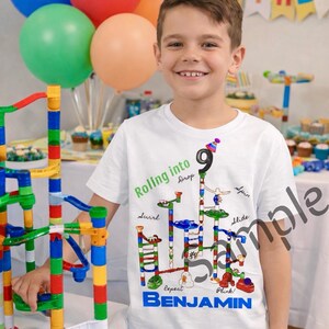 Op de afbeelding: Een persoon draagt een wit t-shirt met een kleurrijk knikkerbaanontwerp en de tekst "Rolling into Drop" en "Benjamin". Het shirt heeft een nummer 9 met een feesthoedje. Kleurrijke ballonnen zijn op de achtergrond.