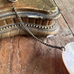 May include: Close-up of a vintage jewelry piece featuring a pearlescent, heart-shaped pendant. The pendant is connected to a delicate chain and a decorative metal clasp. The background includes a gold-colored, ornate object and a wooden surface.