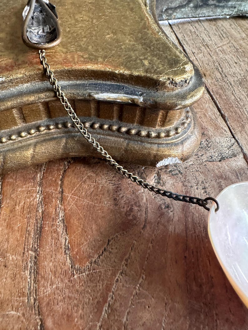 May include: Close-up of a vintage jewelry piece featuring a pearlescent, heart-shaped pendant. The pendant is connected to a delicate chain and a decorative metal clasp. The background includes a gold-colored, ornate object and a wooden surface.