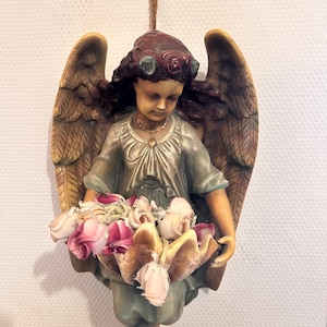 May include: A wall-mounted angel figurine with dark hair, a floral crown, and large feathered wings. The angel wears a muted blue-green gown and holds a large shell-shaped basin filled with a cluster of artificial pink and white roses.