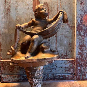 Puede incluir: Un aplique de pared de estilo antiguo con una figura de querubín sosteniendo una tela. El querubín está sentado sobre una base decorativa con un soporte en espiral. La pieza tiene un aspecto envejecido y desgastado, de color marrón dorado cálido.