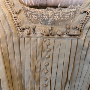 May include: A white cotton blouse with a lace trim and button detailing. The blouse has a pleated front and a V-neck.