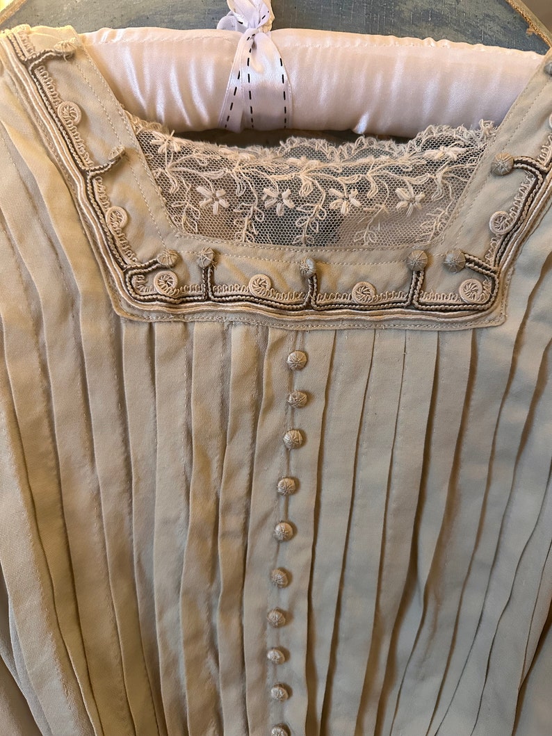May include: A white cotton blouse with a lace trim and button detailing. The blouse has a pleated front and a V-neck.