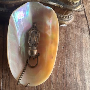 May include: A pearlescent seashell holds a small, antique statue of a religious figure. The statue is attached to a chain and ring. The shell has iridescent colors of pink, yellow, and blue. The background is a wooden surface.