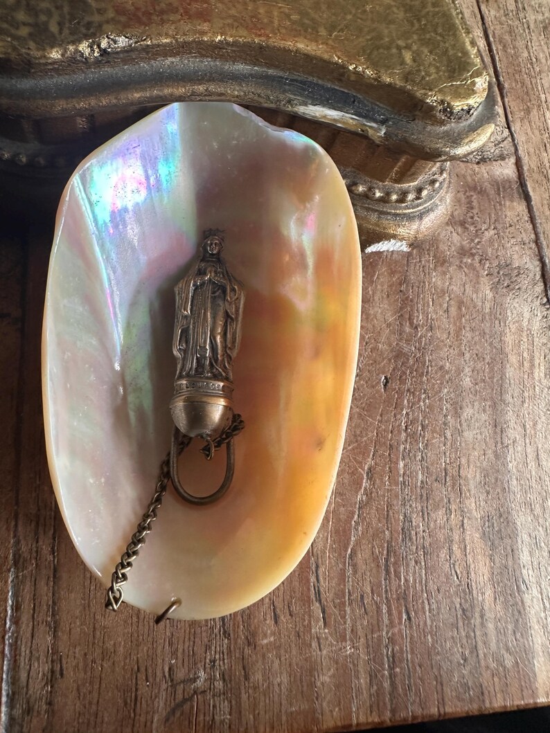 May include: A pearlescent seashell holds a small, antique statue of a religious figure. The statue is attached to a chain and ring. The shell has iridescent colors of pink, yellow, and blue. The background is a wooden surface.