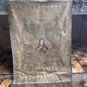 May include: A rectangular, antique textile in a muted gold hue, featuring an embroidered religious design. The central image depicts a figure, angels, and architectural details. The bottom edge is finished with a fringe.