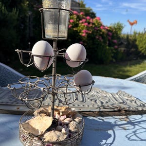 Può includere: Un portauova in metallo vintage con una tazza di vetro sopra. Il supporto ha diversi cestini di filo metallico per le uova ed è decorato con conchiglie. Tre uova bianche sono nei cestini. Il supporto è su un tavolo blu all'aperto.