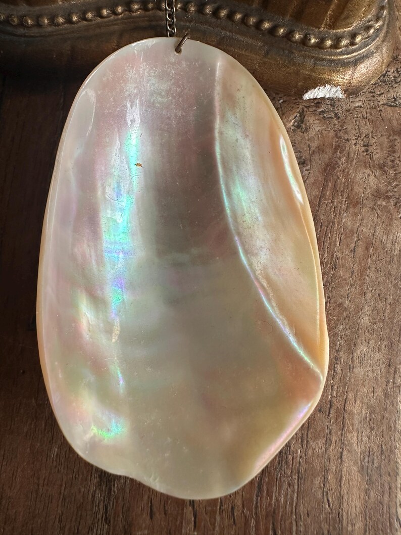 May include: A large, iridescent pendant with a pearlescent sheen. The pendant is a light cream color with hints of pink, blue, and green. It is suspended from a delicate chain. The pendant has a smooth, polished surface.