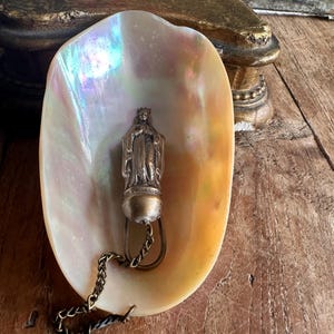 May include: A pearlescent, iridescent seashell holds a small, antique silver-toned religious pendant. The pendant depicts a figure with a crown and is attached to a chain. The shell rests on a weathered wooden surface.