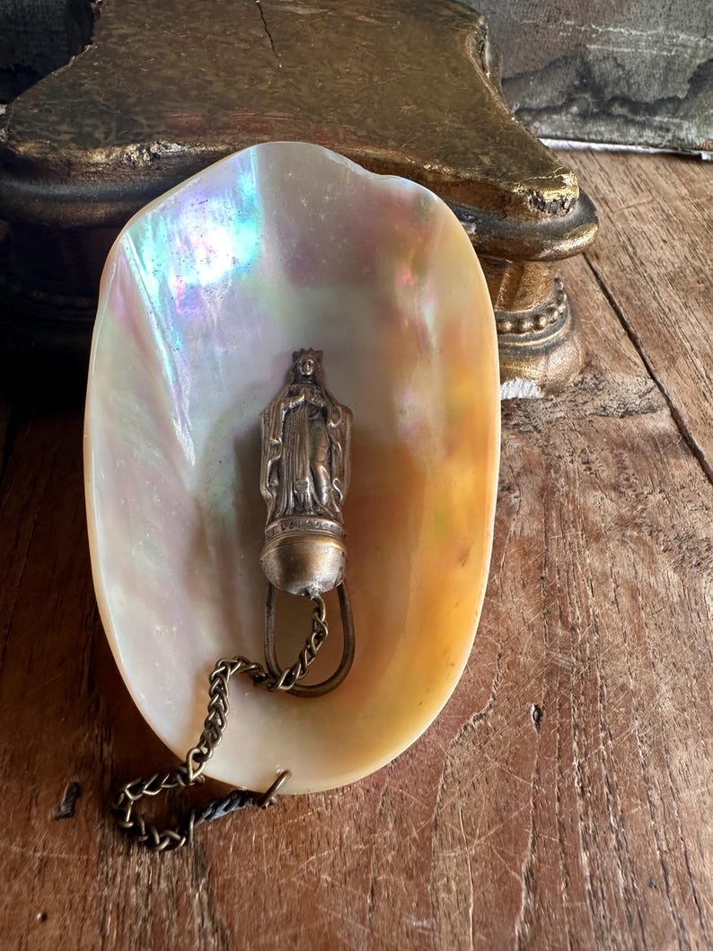 May include: A pearlescent, iridescent seashell holds a small, antique silver-toned religious pendant. The pendant depicts a figure with a crown and is attached to a chain. The shell rests on a weathered wooden surface.