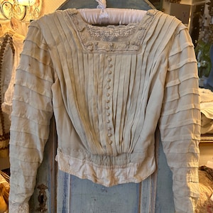 May include: A vintage white blouse with a lace trim and button fastenings. The blouse has a pleated bodice and long sleeves with ruched detailing.