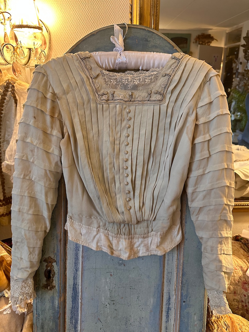 May include: A vintage white blouse with a lace trim and button fastenings. The blouse has a pleated bodice and long sleeves with ruched detailing.