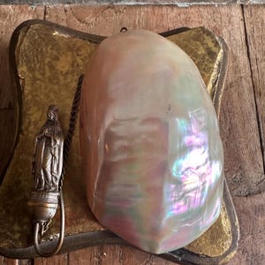 May include: A large, iridescent, pearlescent shell rests on a gold-toned, decorative base. A small, detailed metal religious figure hangs from a chain. The shell displays a range of colors, including pink, purple, and green.