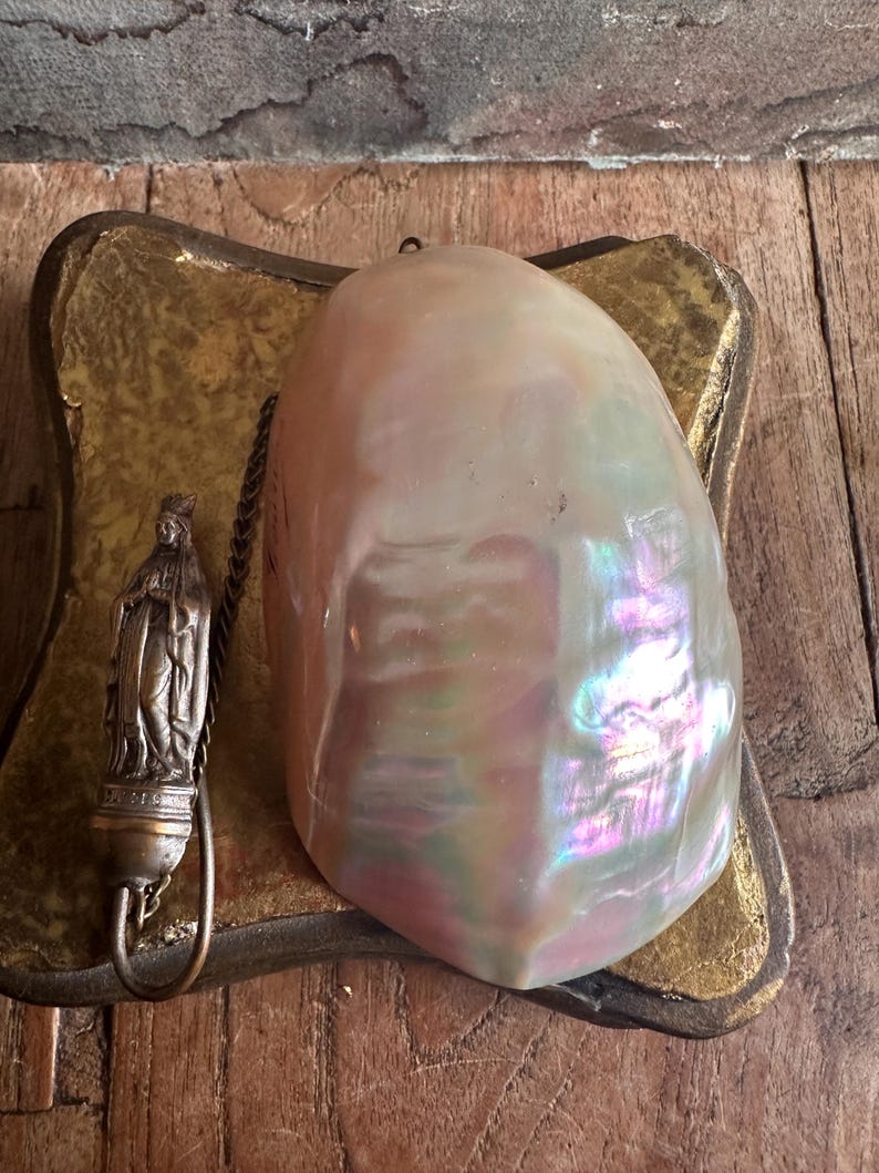 May include: A large, iridescent, pearlescent shell rests on a gold-toned, decorative base. A small, detailed metal religious figure hangs from a chain. The shell displays a range of colors, including pink, purple, and green.