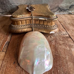 May include: A pearlescent, iridescent shell-shaped pendant hangs from a gold-toned chain. The pendant is suspended in front of a gold-painted, ornate, rectangular base. The background is a weathered wooden surface.