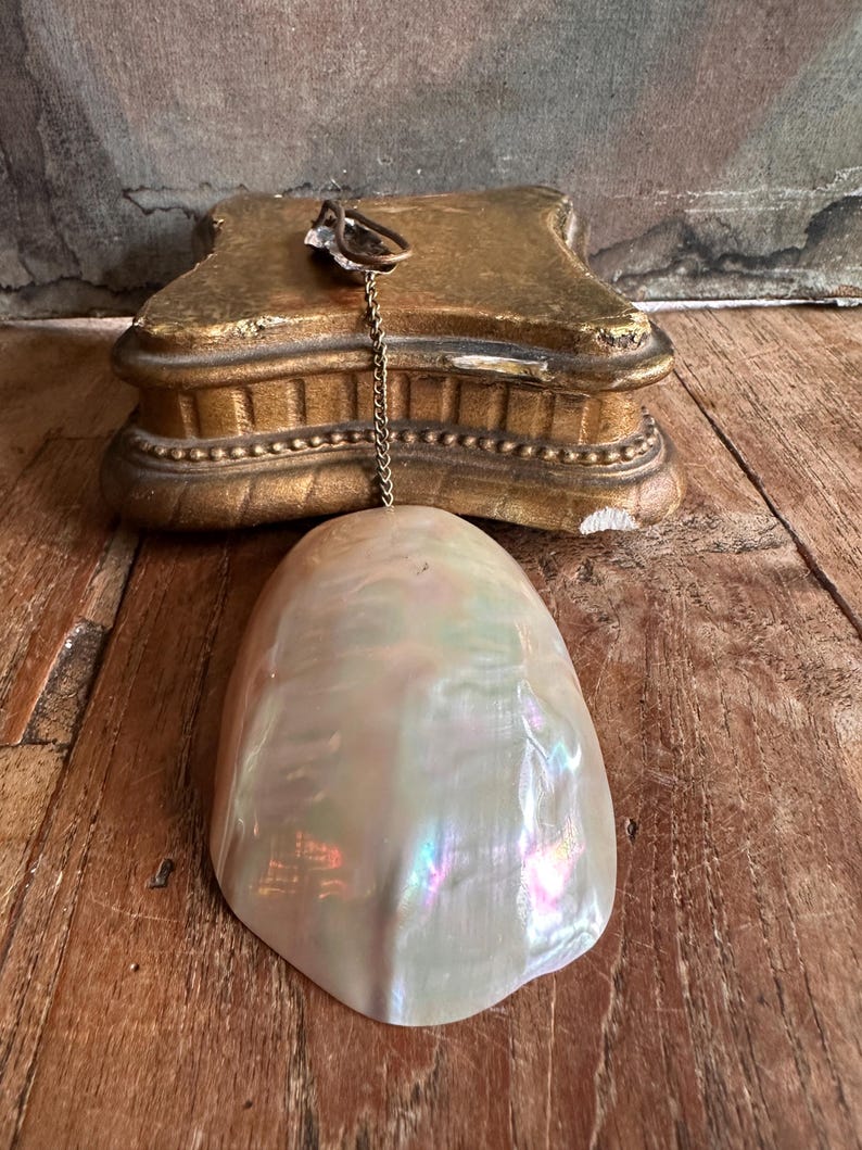 May include: A pearlescent, iridescent shell-shaped pendant hangs from a gold-toned chain. The pendant is suspended in front of a gold-painted, ornate, rectangular base. The background is a weathered wooden surface.