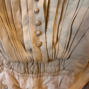 May include: Close-up of a beige garment with a gathered waist and a row of decorative buttons. The fabric is pleated and has a slightly textured surface.