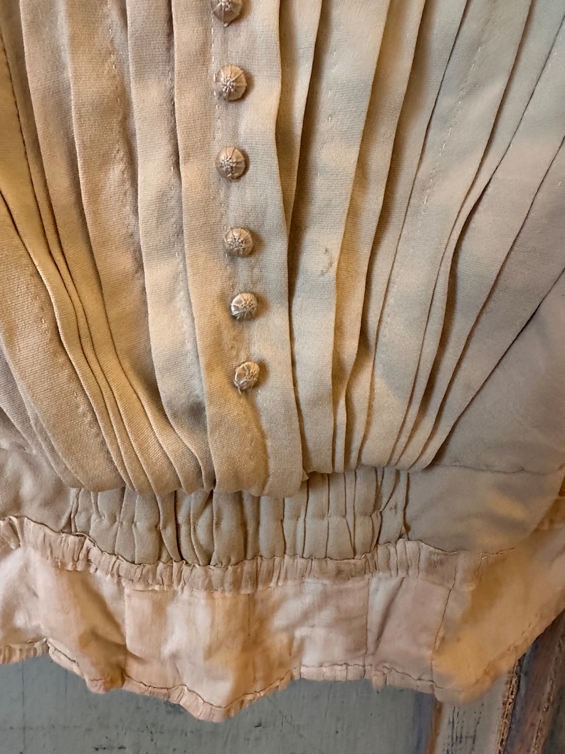 May include: Close-up of a beige garment with a gathered waist and a row of decorative buttons. The fabric is pleated and has a slightly textured surface.