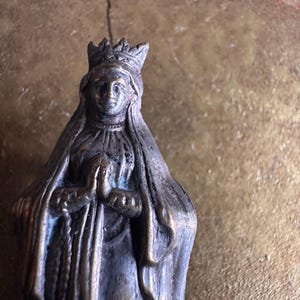 May include: A small, antique-style statue of a woman wearing a crown and draped in a veil. The statue is made of a dark metal with visible wear and tear, suggesting age. The woman's hands are clasped in prayer.