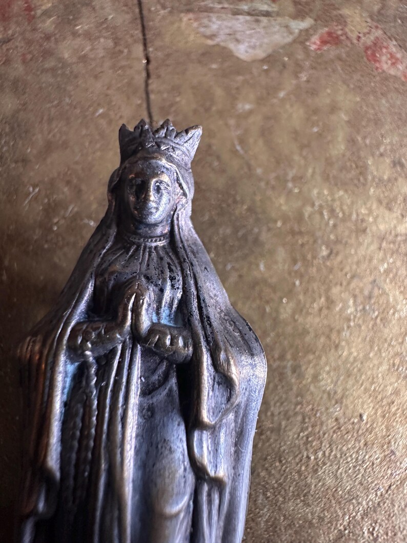 May include: A small, antique-style statue of a woman wearing a crown and draped in a veil. The statue is made of a dark metal with visible wear and tear, suggesting age. The woman's hands are clasped in prayer.