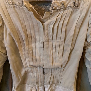 May include: A vintage white cotton blouse with a pleated front and lace trim around the neckline. The blouse has a button-up closure and long sleeves.