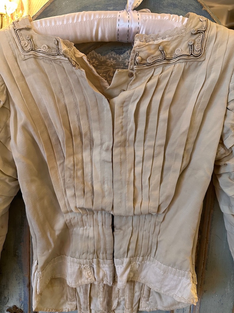 May include: A vintage white cotton blouse with a pleated front and lace trim around the neckline. The blouse has a button-up closure and long sleeves.