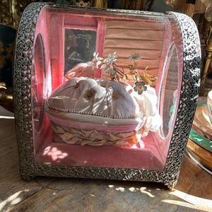 May include: A vintage glass display case with a pink velvet interior. The case features a delicate white and gold floral arrangement and a small pink and white pillow.