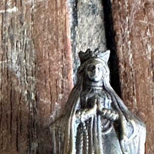 May include: A small, antique-style statue of a robed figure wearing a crown. The statue is a muted bronze color and features intricate details, including a rosary. The background is a weathered wooden surface.