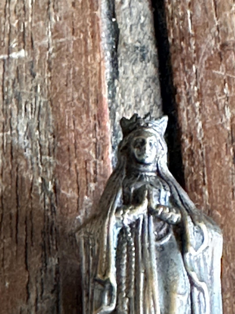 May include: A small, antique-style statue of a robed figure wearing a crown. The statue is a muted bronze color and features intricate details, including a rosary. The background is a weathered wooden surface.