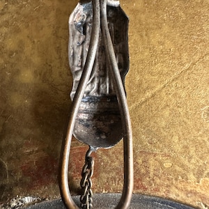May include: Antique silver-toned religious pendant with a detailed figure and a teardrop-shaped frame. A small chain hangs from the bottom. The pendant is displayed against a gold-toned background.