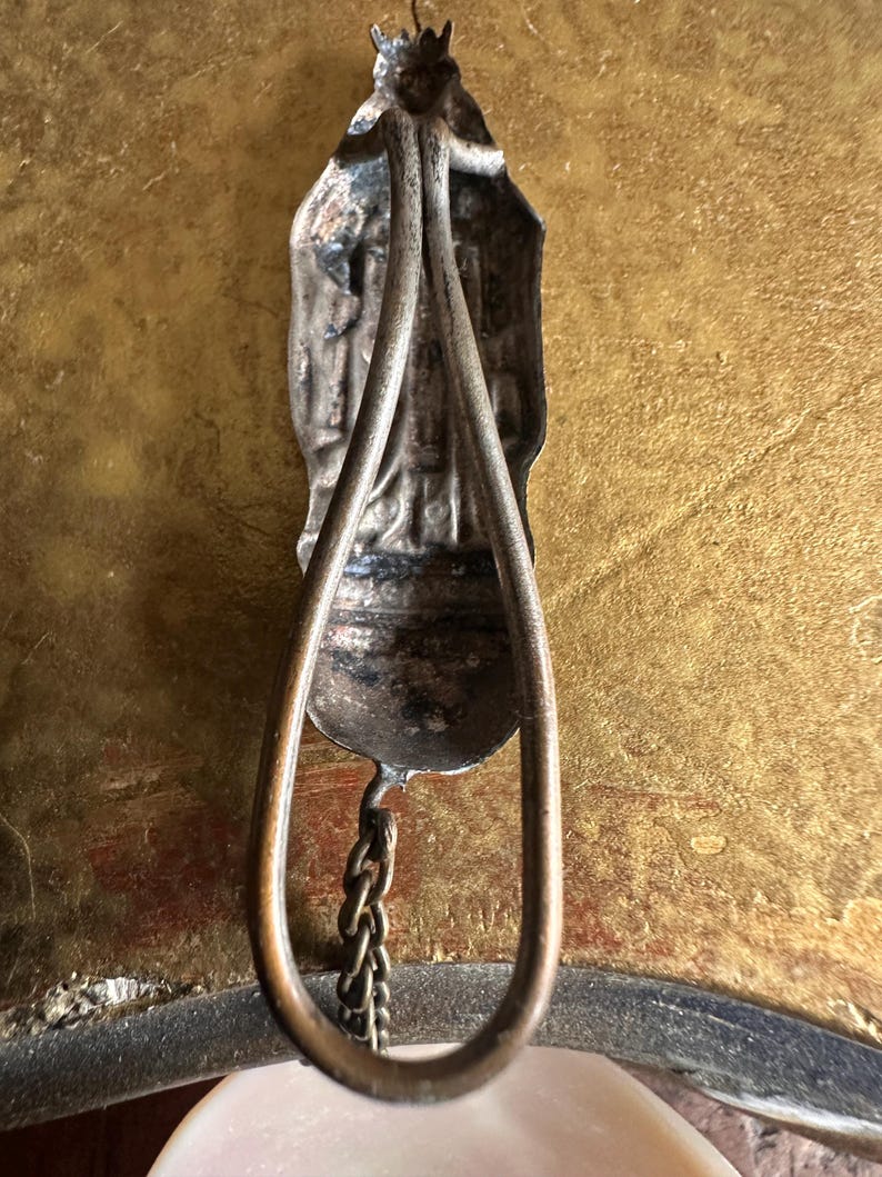 May include: Antique silver-toned religious pendant with a detailed figure and a teardrop-shaped frame. A small chain hangs from the bottom. The pendant is displayed against a gold-toned background.
