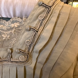May include: A close-up of the neckline of a beige garment with a decorative trim of white and black embroidery. The trim features a series of small, round buttons.