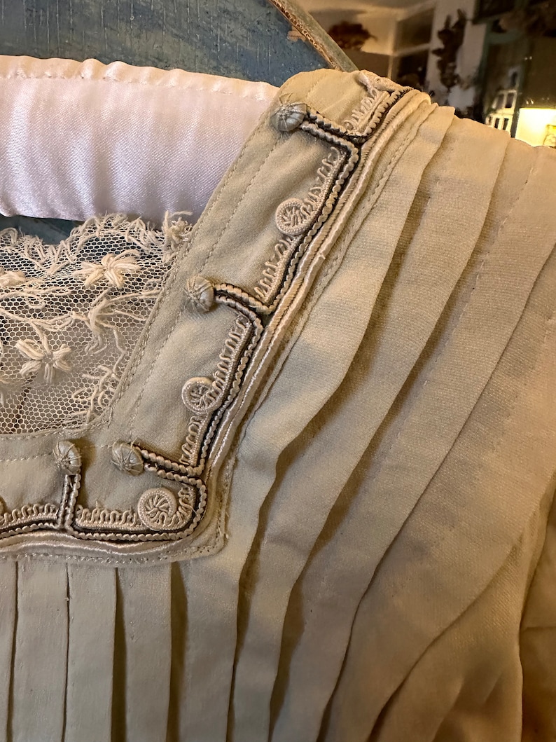 May include: A close-up of the neckline of a beige garment with a decorative trim of white and black embroidery. The trim features a series of small, round buttons.