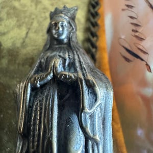 May include: A detailed, antique-style statue of a woman wearing a crown and draped in a long cloak. The statue is made of a metallic material with a weathered appearance. The woman's hands are clasped in prayer. The background is blurred, with hints of gold and other colors.