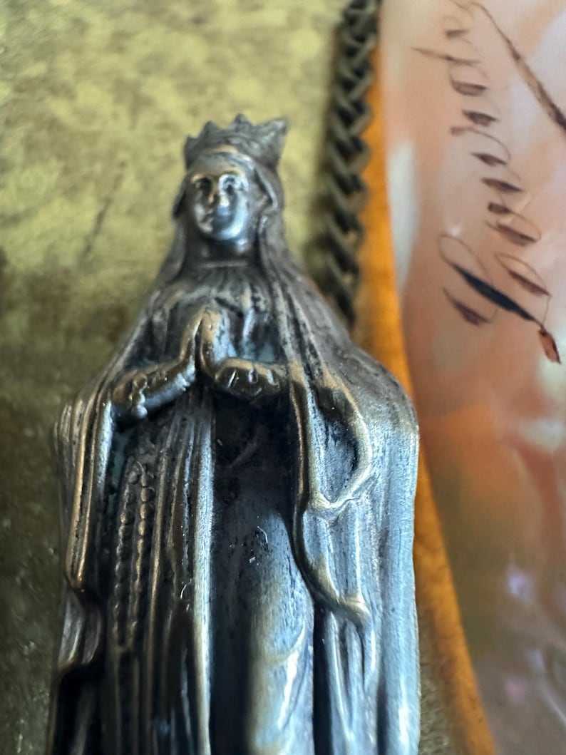 May include: A detailed, antique-style statue of a woman wearing a crown and draped in a long cloak. The statue is made of a metallic material with a weathered appearance. The woman's hands are clasped in prayer. The background is blurred, with hints of gold and other colors.