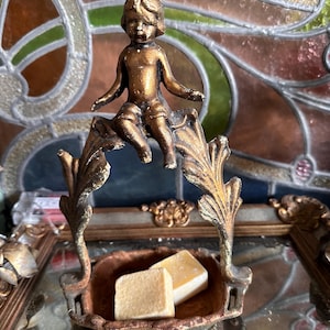 Może przedstawiać: Złota metalowa miska z figurką cheruba na górze. Miska ma ozdobny, bogato zdobiony wzór i zawiera dwa małe, kwadratowe, białe kawałki mydła.