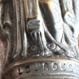 May include: Close-up of a silver-toned religious statue base. The word "LOURDES" is engraved on the base. The statue appears to be of a saint or religious figure.