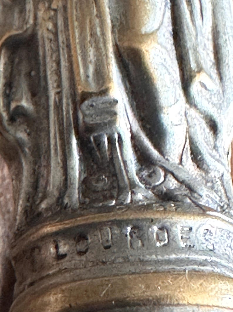 May include: Close-up of a silver-toned religious statue base. The word "LOURDES" is engraved on the base. The statue appears to be of a saint or religious figure.