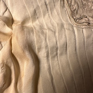 May include: A close-up of a white, pleated fabric with a decorative trim along the edge. The fabric has a subtle sheen and a slightly textured surface.