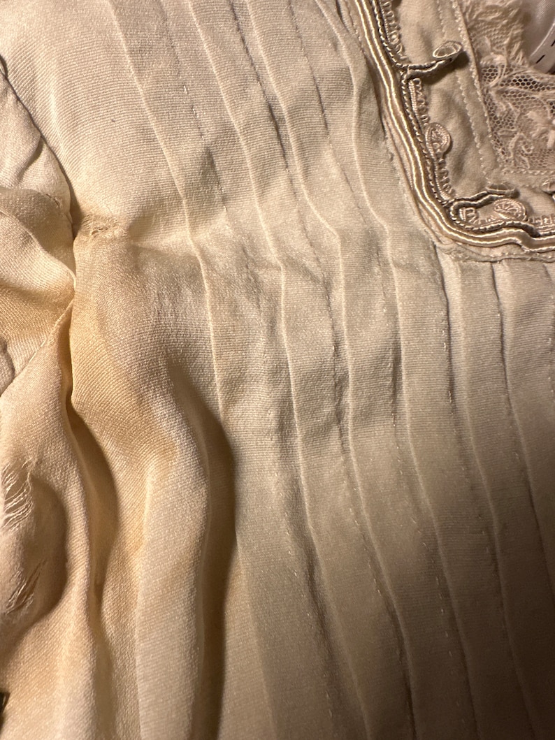 May include: A close-up of a white, pleated fabric with a decorative trim along the edge. The fabric has a subtle sheen and a slightly textured surface.