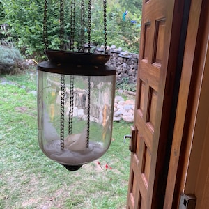 May include: A glass candle lantern with a white candle inside, hanging from a chain. The lantern is suspended in front of a wooden door with a paneled design.