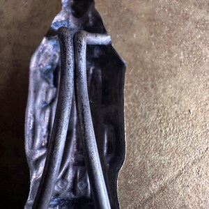 May include: A vintage silver-toned religious figurine with a crown, holding two metal rods. The figure has intricate details and is set against a textured, golden-brown background. The rods are curved and appear to be part of the figure's design.