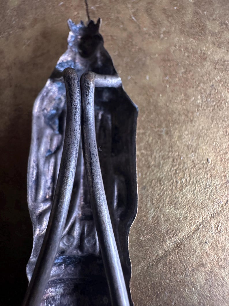 May include: A vintage silver-toned religious figurine with a crown, holding two metal rods. The figure has intricate details and is set against a textured, golden-brown background. The rods are curved and appear to be part of the figure's design.