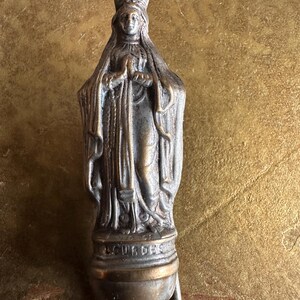 May include: A bronze-toned religious pendant featuring a detailed statue of a robed figure with a crown, hands clasped in prayer. The word "LOURDES" is inscribed on the base. The pendant has a small, round base with a loop for attachment.