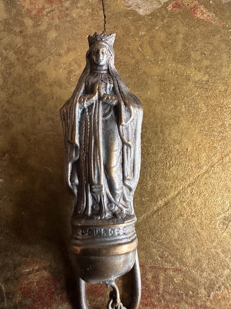 May include: A bronze-toned religious pendant featuring a detailed statue of a robed figure with a crown, hands clasped in prayer. The word "LOURDES" is inscribed on the base. The pendant has a small, round base with a loop for attachment.