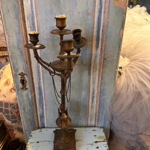 May include: A vintage, ornate, gold-toned metal candelabra with four candle holders. The candelabra has a decorative, bird-like base and a chain hanging from the centre.