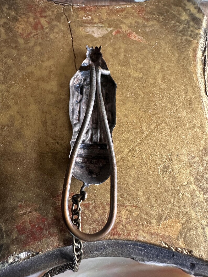 May include: Antique silver-toned religious pendant with a figure wearing a crown and cape. The pendant has a teardrop-shaped handle and a small chain. The background is a textured gold color.