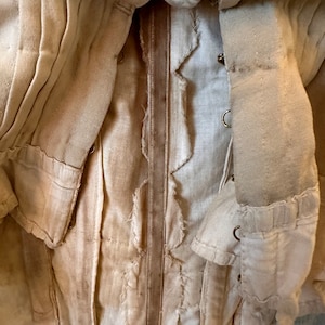 May include: A close-up of a white garment with a brown leather strap. The garment is made of a thick, woven fabric and has a frayed edge. The strap is sewn to the garment and has a decorative stitch.